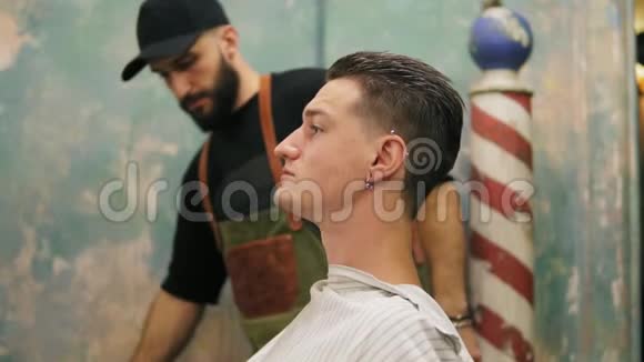 年轻的胡子理发师从时尚的客户脱下沙龙披风他的耳朵和塔图满意视频的预览图