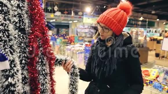 漂亮的年轻女人戴着眼镜和红帽子选择圣诞装饰作为圣诞树买家找到了便宜的视频的预览图