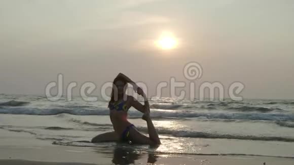美丽苗条的女孩穿着泳衣在日落时在海滩上做瑜伽4K视频的预览图