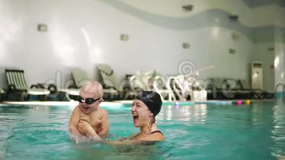 年轻的母亲戴着防护眼镜抱着她的小男孩教她如何在游泳池里游泳宝宝踢脚视频的预览图