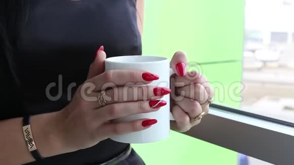 迷人的年轻女子站在窗户附近喝着白色杯子里的茶或咖啡视频的预览图