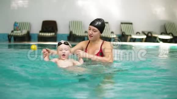 年轻的妈妈正在教她可爱的小儿子在游泳池里游泳她牵着他的手在玩视频的预览图