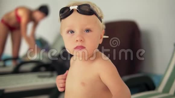 一个戴着护目镜的漂亮孩子坐在泳池边看着镜头等待母亲的画像视频的预览图