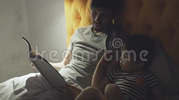 年轻的父亲和小儿子躺在家里的床上晚上睡觉前读童话书视频的预览图