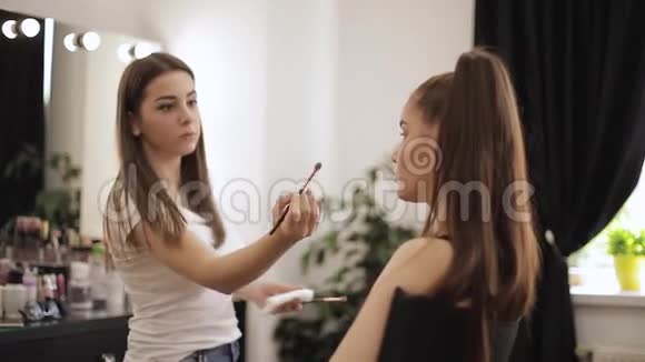 一位美丽的女性化妆师为一个年轻的红发女孩化妆她坐在一面大镜子前的美容院里视频的预览图