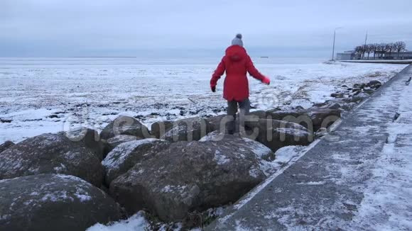 穿着冬装的女孩在海湾的白雪覆盖的岩石上跳跃视频的预览图
