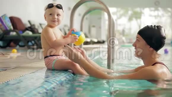 年轻的母亲正在教她可爱的金发幼儿在游泳池里游泳她坐在水边视频的预览图