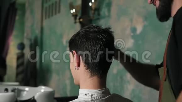 一位坐在理发店椅子上的男客户身穿围裙梳头的胡子理发师慢镜头旋转视频的预览图