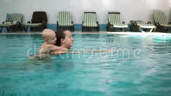 年轻的母亲正在游泳池里游泳背着她可爱的小蹒跚学步的孩子可爱的小男孩拥抱着他视频的预览图