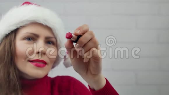 圣诞快乐圣诞老人女孩在玻璃字上写口红圣诞快乐戴帽子的漂亮女孩发信息视频的预览图