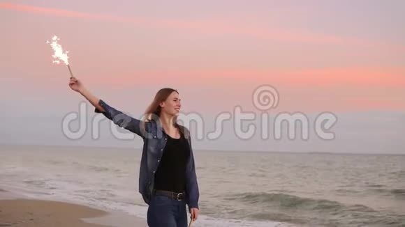 日落时分年轻迷人的女人站在海边手里拿着燃烧的闪闪发光的蜡烛慢镜头视频的预览图