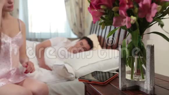 美丽的妻子在平板电脑上看照片丈夫睡在床上视频的预览图