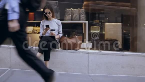 年轻漂亮的女商人坐在商店橱窗边喝着咖啡休息时用着智能手机视频的预览图