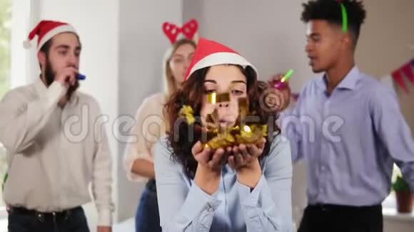 年轻貌美的女人戴着圣诞帽从手中吹出金色的纸屑而她的同事在聚会视频的预览图