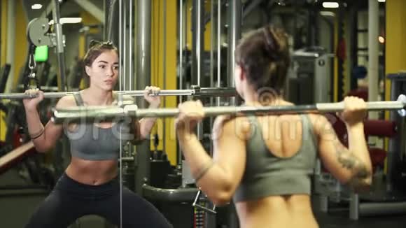 年轻的女运动员在镜子前的健身房里用杠铃做蹲视频的预览图