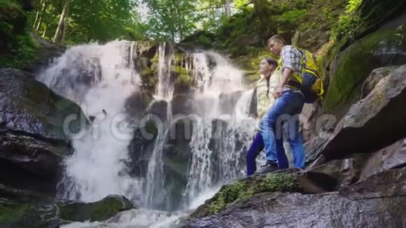 一对年轻的游客背着背包欣赏着山里的瀑布站在光滑的石头上视频的预览图