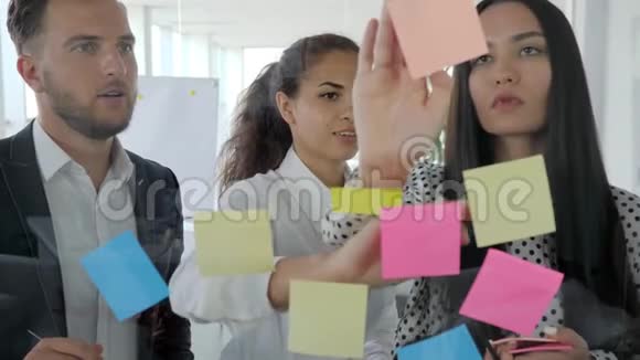 年轻的商业团队在贴在玻璃墙上的黏糊糊的纸条上写下想法办公室里的人看着五颜六色的东西视频的预览图