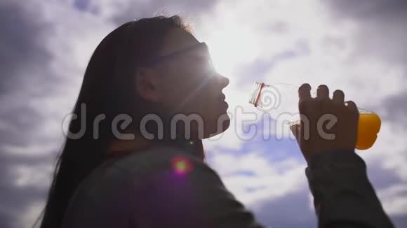 年轻漂亮的女人在夏天公园的户外天空背景上喝橙汁慢动作视频的预览图