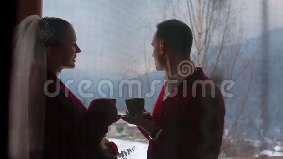 新郎和新娘在村里木木木屋的阳台上用杯子喝咖啡或茶然后用雪吻视频的预览图