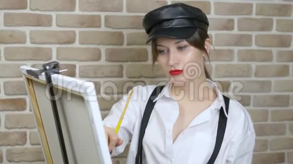 戴皮帽的女艺术家在画布上靠砖墙画一幅画视频的预览图