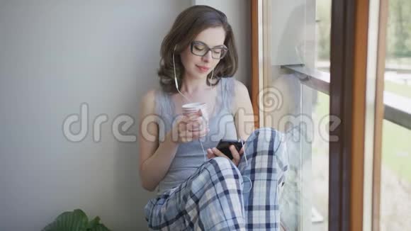 年轻女子坐在窗前用智能手机带耳机视频的预览图