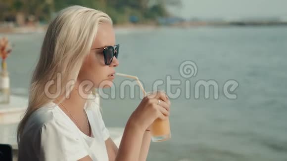年轻迷人的女孩在海滩上喝健康的果汁芒果橙的金发肖像视频的预览图