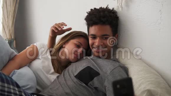 年轻美丽有爱心的多民族夫妇在智能手机摄像头上自拍躺在床上视频的预览图
