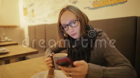 快乐惊讶的女人使用智能手机坐在咖啡馆里年轻的白种人女孩戴着眼镜打短信和喝咖啡视频的预览图