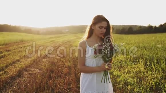 一个穿着白色连衣裙的女孩站在草地上夕阳下带着一束野花看着镜头视频的预览图