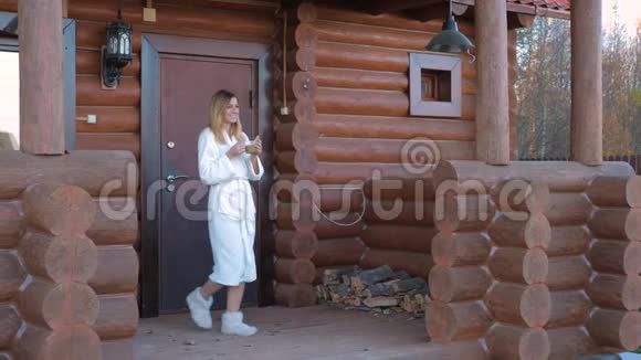 穿着白色浴衣和拖鞋的女人带着咖啡走进门廊和木屋视频的预览图