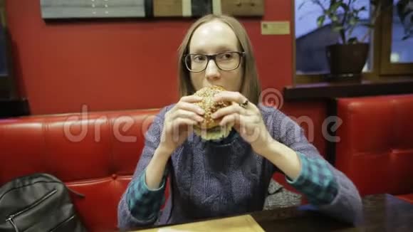 戴眼镜的年轻女人在咖啡馆里吃快餐汉堡包和炸薯条可爱的学生女孩坐在视频的预览图