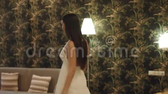 穿着白色连衣裙和高跟鞋留着长发的布鲁内特走到全景窗前拉开窗帘视频的预览图