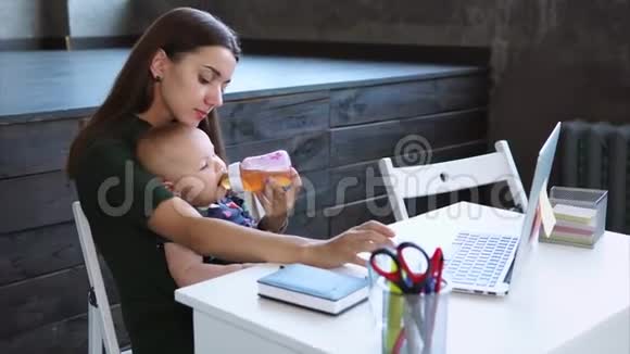 年轻的母亲手里拿着她的孩子坐在桌子上用笔记本视频的预览图