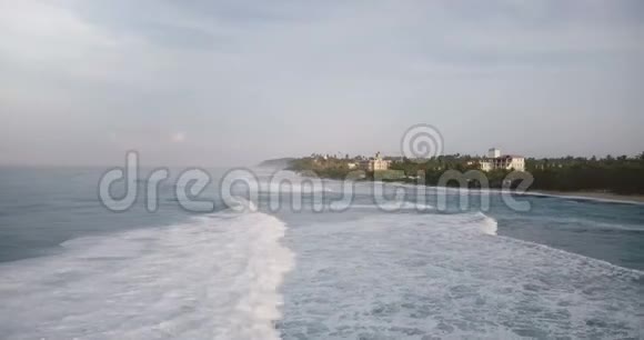 无人机在波涛汹涌的海浪上翻滚到达风景如画的度假胜地海岸那里有树木和房屋视频的预览图