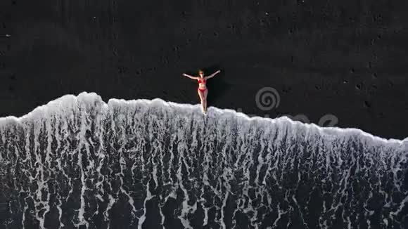 一个穿红色泳衣的女孩躺在冲浪线上的黑色海滩上的俯视图加那利特内利岛海岸视频的预览图
