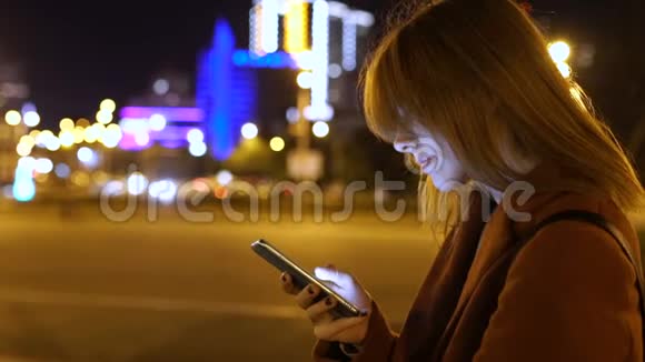 迷人的年轻女性在夜间在街上散步时使用智能手机慢动作视频的预览图