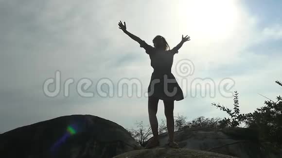 站在岩石上的年轻快乐女人的剪影通过太阳举起双手顶着天空慢动作视频的预览图