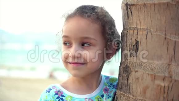 海滩上快乐的小女孩穿着一件蓝色的连衣裙戴着一棵棕榈树看着相机微笑着有点害羞儿童视频的预览图