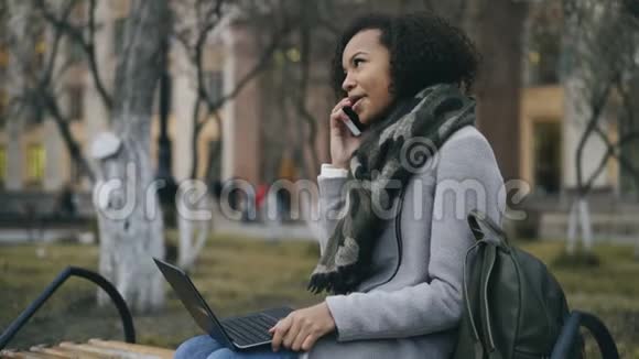 漂亮的非裔美国学生女孩在户外坐在长凳上用笔记本电脑说话视频的预览图