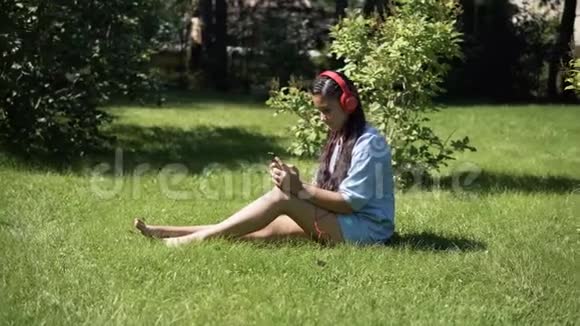 小女孩听音乐戴着耳机坐在公园的草地上4K视频的预览图