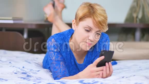 美丽的女人穿着蓝色连衣裙躺在床上用智能手机视频的预览图