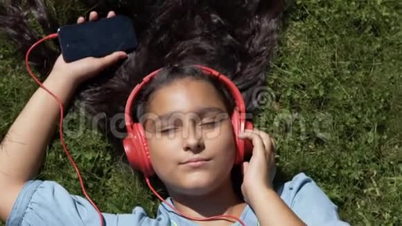 长着黑色长发的漂亮女孩躺在公园的草地上用红色耳机听音乐视频的预览图