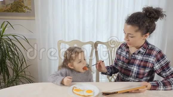 女人用香肠喂女儿可爱的小女孩用平板电脑看动画片视频的预览图