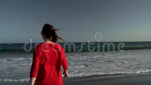 幸福的年轻女子穿着红色的连衣裙在日落时步行到海边的水上无忧无虑的现代生活理念慢慢视频的预览图