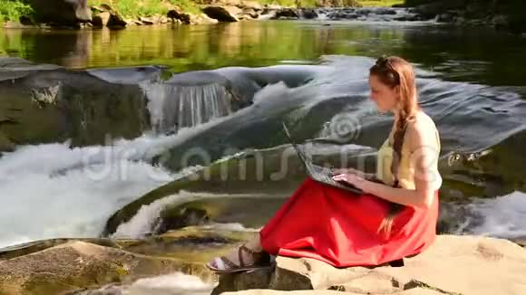 美丽的长发女孩穿着红色裙子手提电脑坐在山河瀑布背景上的岩石上自由职业概念视频的预览图