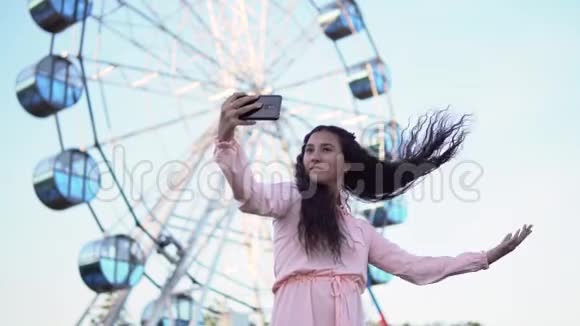 一个穿着连衣裙留着长发的女孩用智能手机在摩天轮附近自拍慢动作视频的预览图