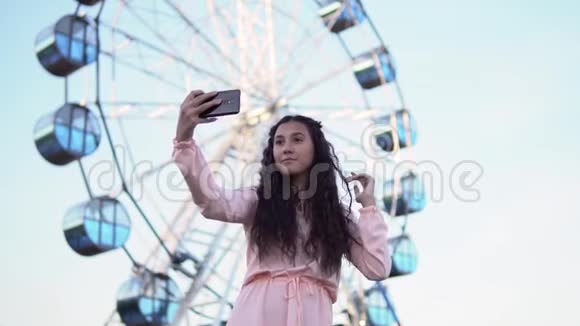 一个穿着连衣裙留着长发的年轻女孩用一部智能手机在摩天轮附近自拍慢动作视频的预览图