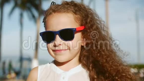 一个快乐可爱的小女孩的特写肖像她卷发和红色太阳镜对着镜头视频的预览图