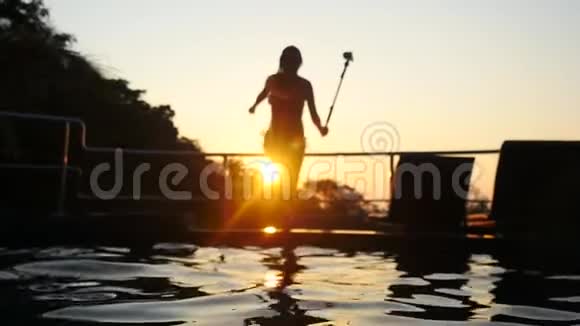 美丽的年轻女子在美丽的夕阳下在游泳池里跳着剪影镜头闪耀视频的预览图
