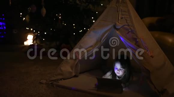 小女孩坐在家里的帐篷里背景是一棵圣诞树在电子平板电脑上玩游戏视频的预览图